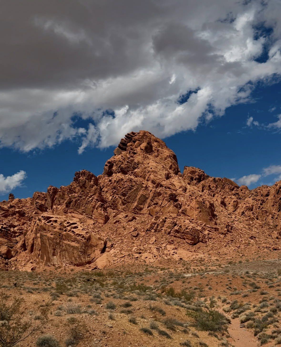 Jagged red sandstone peak rising above desert scrub and white clouds