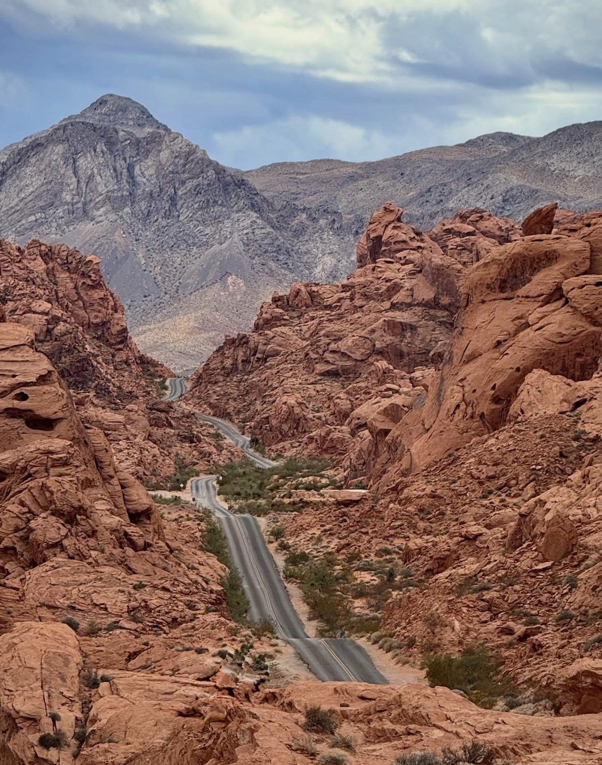 Road dipping between red rocks toward grey mountains, cloudy afternoon